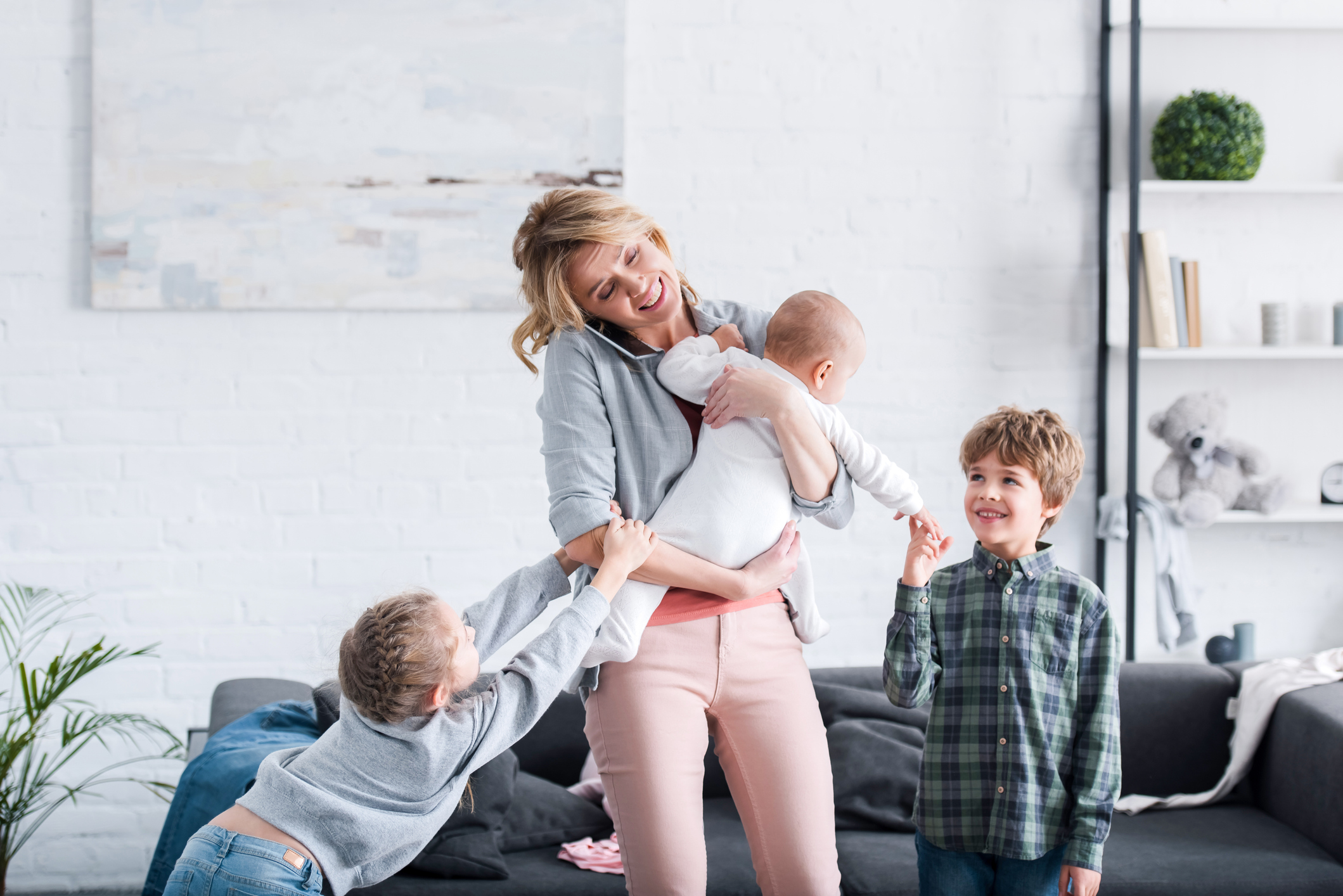 exhausted mother talking by smartphone and holding infant child while naughty siblings playing in room