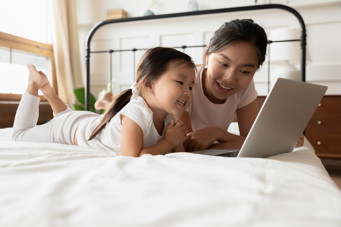Asian mother and little daughter lying in bed with laptop